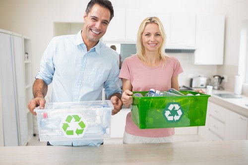 A smiling man and woman standing in a modern kitchen holding clear and green recycling bins with white recycling symbols, indicating waste segregation. The man, wearing a light blue shirt, holds the clear bin filled with recyclable plastic bottles and containers, positioned on his right. The woman, dressed in a pink top, holds a green bin containing assorted recyclable items, placed on her left. The background features white kitchen cabinets, a stainless steel sink, and various small appliances, with natural light illuminating the space. The scene reflects environmentally conscious waste management practices, consistent with rubbish removal services offered by House Clearance High Wycombe, situated within the local area of High Wycombe, Buckinghamshire, and the surrounding region, subtly supporting local SEO relevance.