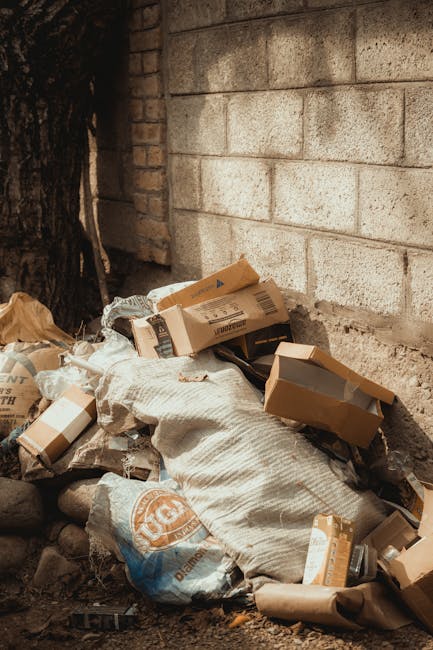 Two large black plastic bags filled with waste material, placed on the sidewalk near a wooden fence and a lamppost in an outdoor urban environment. The bags appear crumpled and slightly torn, indicati