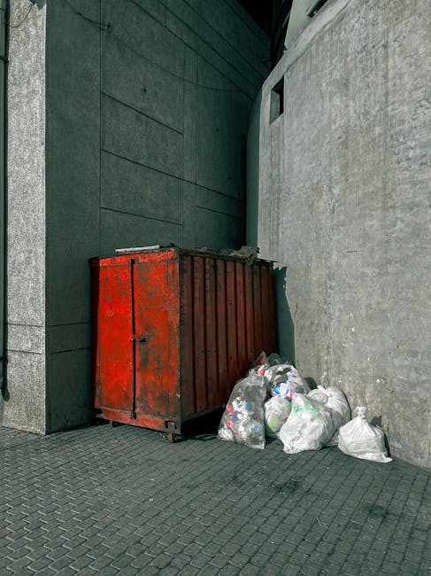 A weathered red metal rubbish skip positioned in a corner between a dark green wall and a light grey concrete wall, with several white and clear plastic rubbish bags stacked beside it on a paved surfa