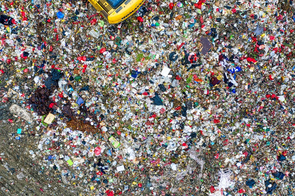 A weathered red metal rubbish skip positioned in a corner between a dark green wall and a light grey concrete wall, with several white and clear plastic rubbish bags stacked beside it on a paved surfa